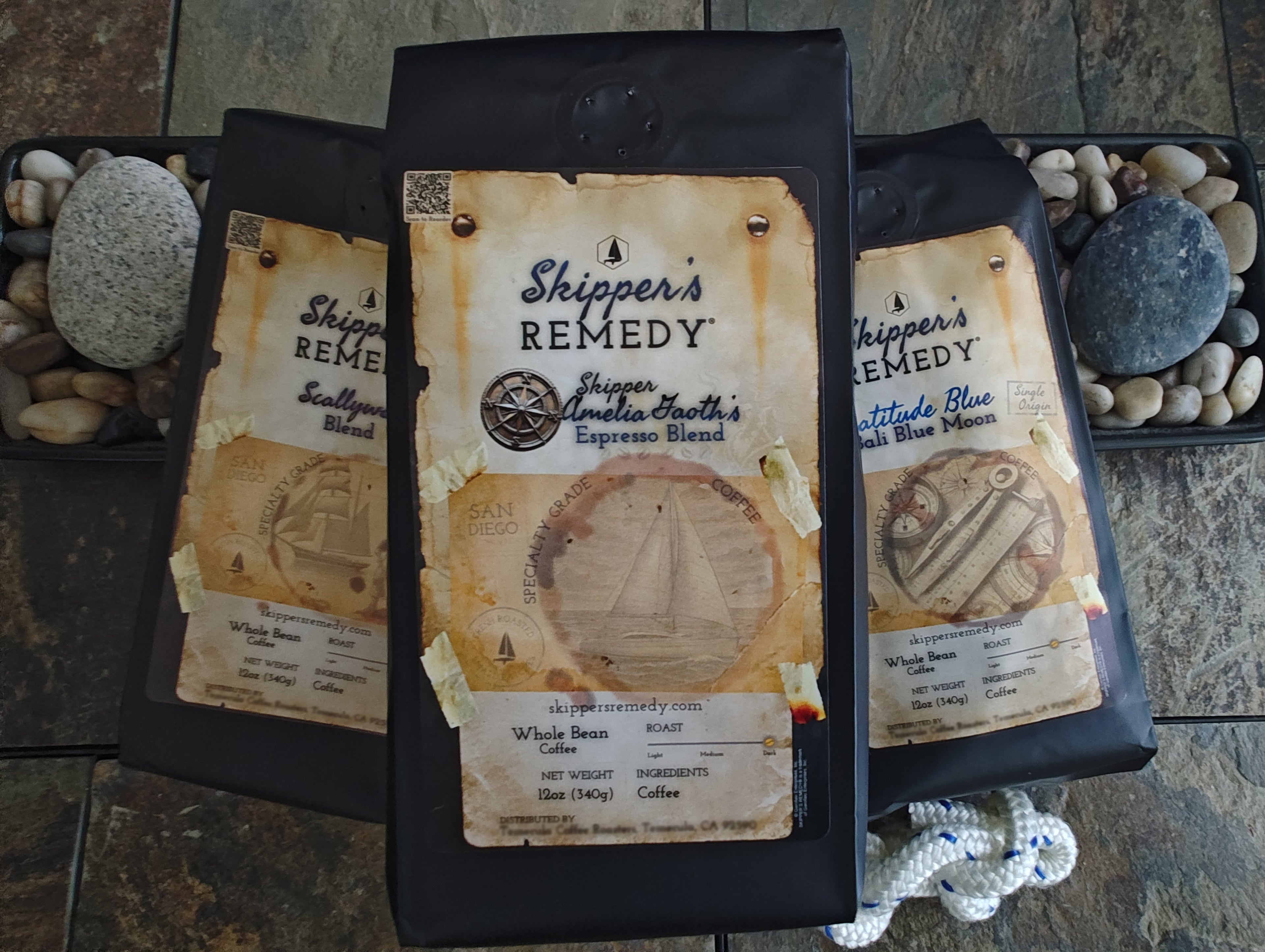 Three packages of 'Skipper's Remedy' coffee (Skipper Amelia Gaoth's Espresso Blend, Latitude Blue - Bali Blue Moon, and Scallywag Blend) with decorative labels on a stone surface.