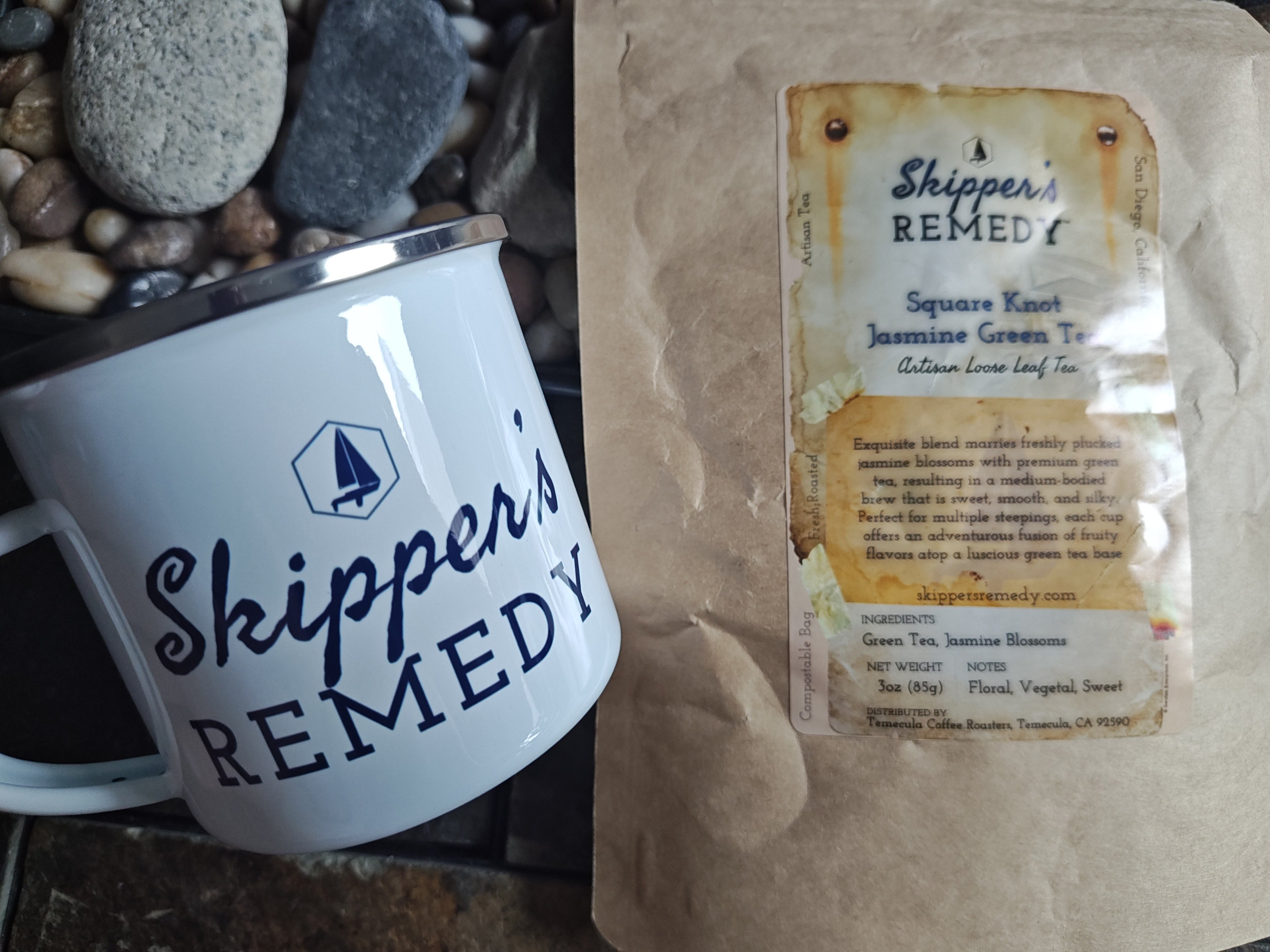 White mug with 'Skipper's Remedy' branding next to a package of Skipper's Remedy - Square Knot Jasmine Green Tea, on a stone surface.