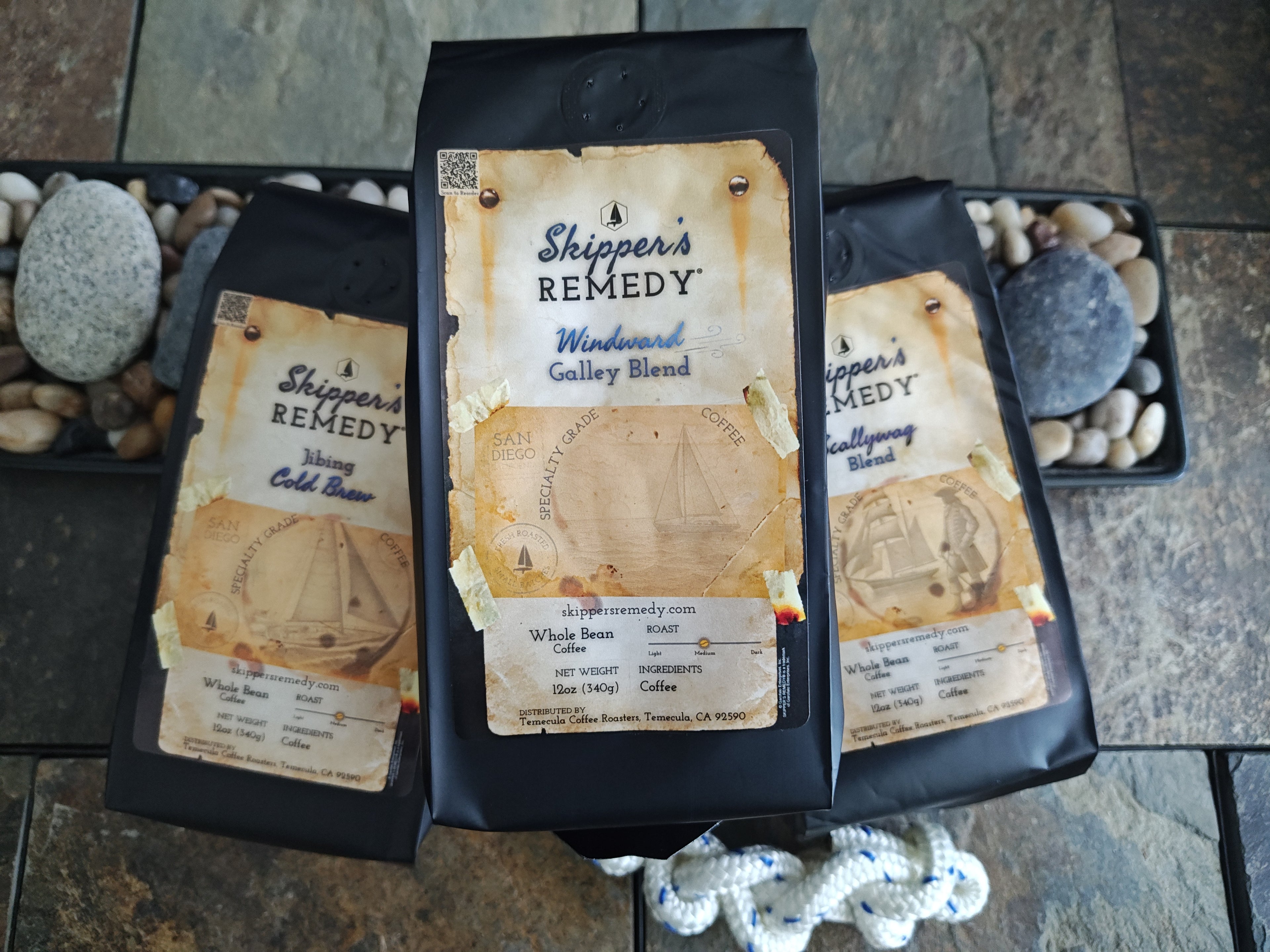Three packages of 'Shipper's Remedy' coffee (Windward Galley Blend, Jibing Cold Brew, Scallywag Blend) on a stone surface.