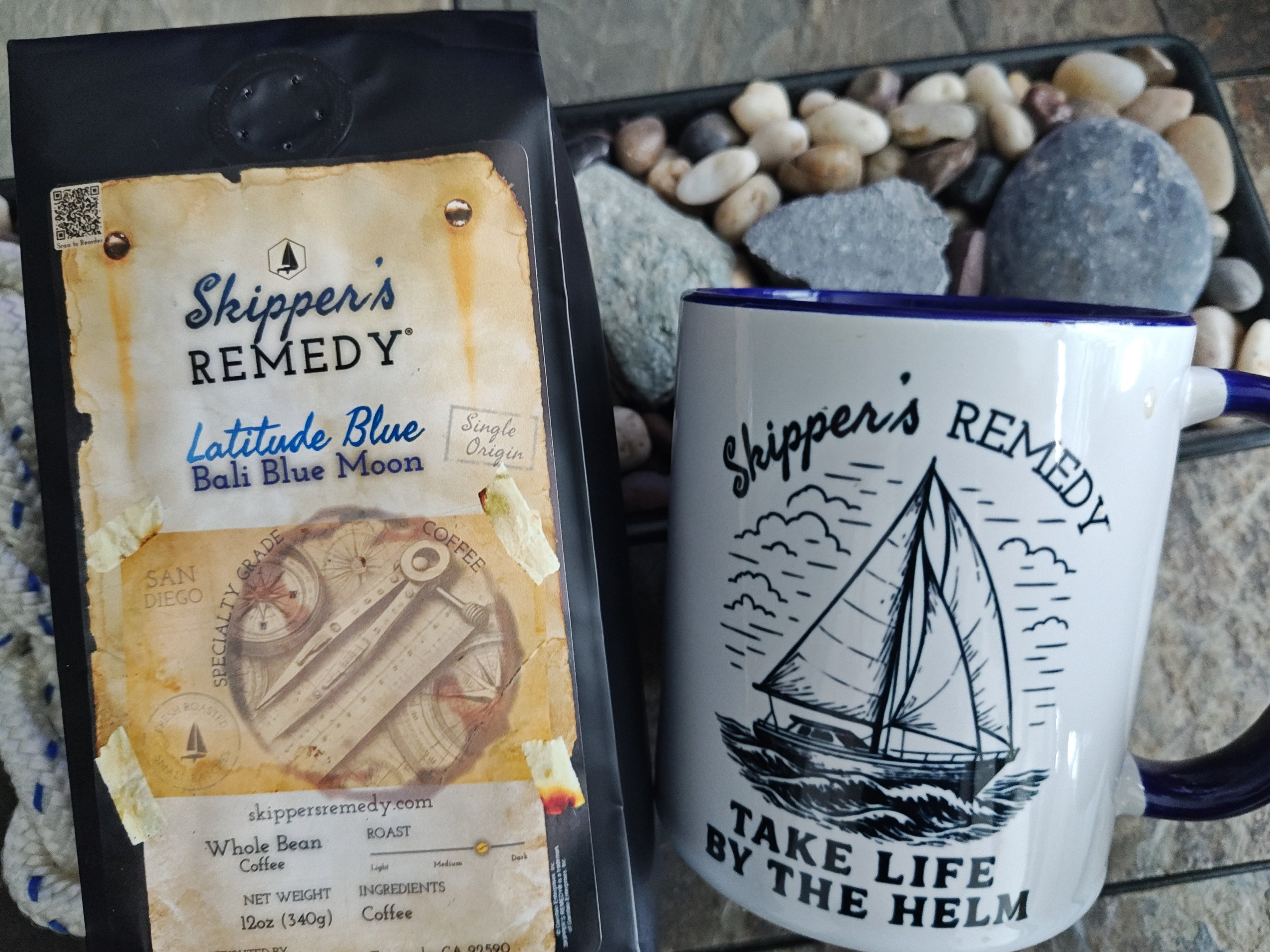 Mug with text and design on a stone surface, next to a branded coffee mug "Skipper's Remedy - Take Life By the Helm".