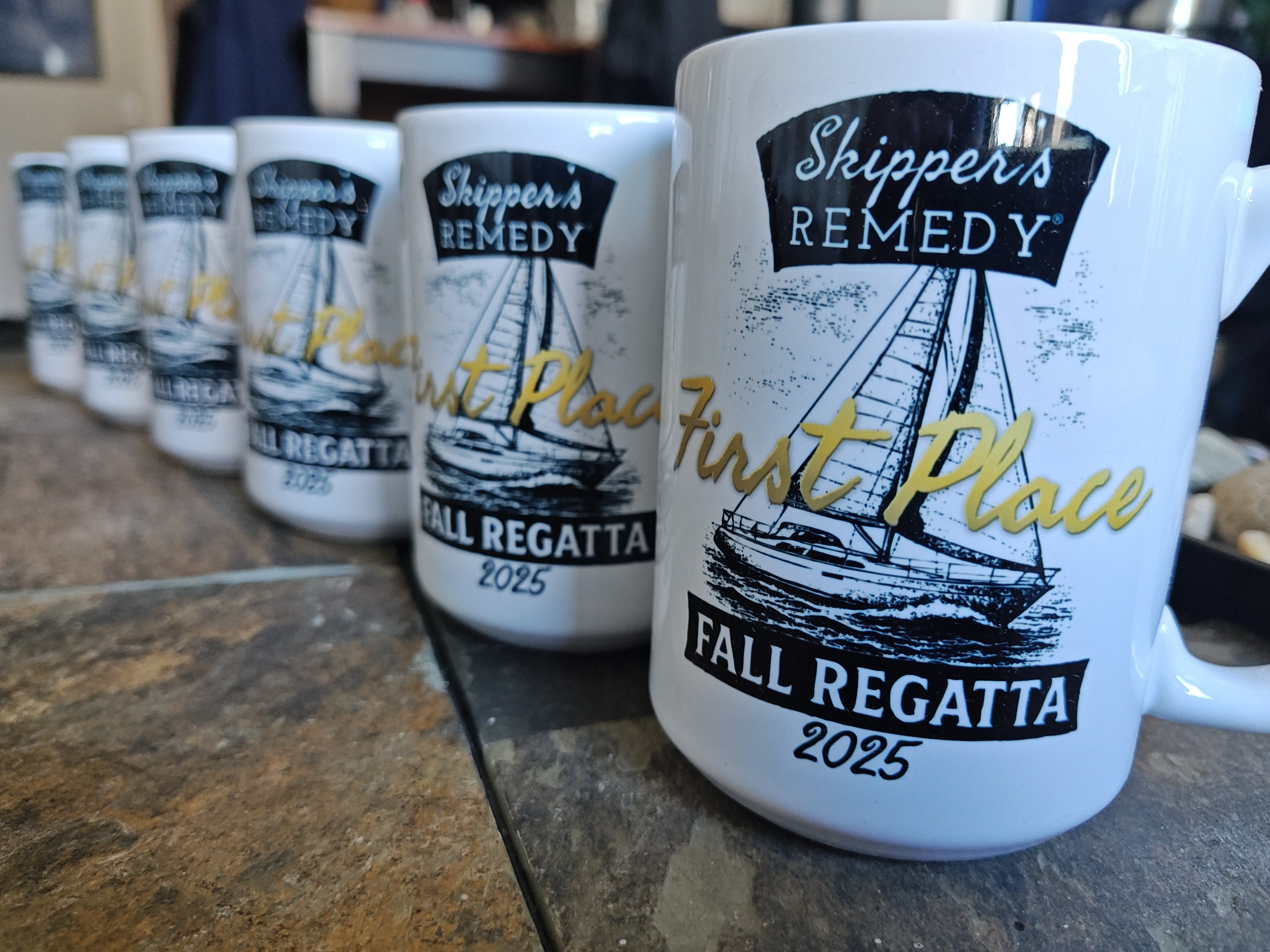 Row of white mugs with 'Skipper's Remedy' branding and sailing design on a stone surface.
